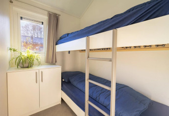 Gezellige lodgekamer met stapelbedden, blauw beddengoed, witte kast, plant en raam met gordijn.