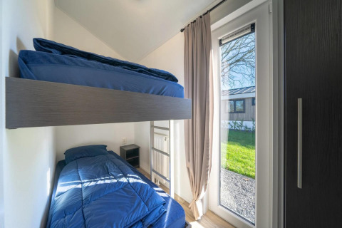Chambre moderne en lodge avec lits superposés, linge de lit bleu et grande fenêtre donnant sur le jardin.