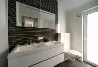 Modernes Badezimmer mit Doppelwaschbecken, großem Spiegel, Wandtoilette und viel Tageslicht.