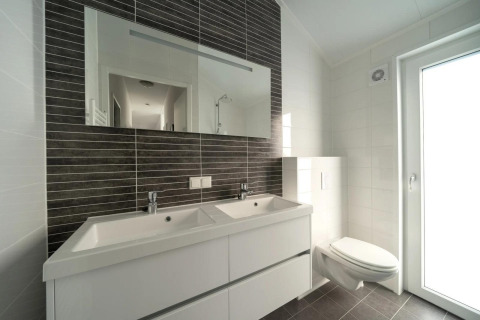 Baño moderno con doble lavabo, espejo grande, inodoro suspendido y abundante luz natural de la puerta.