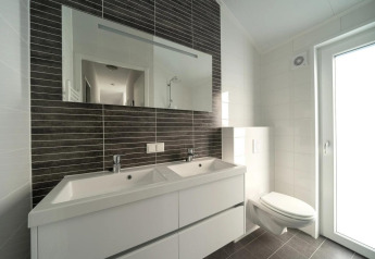Moderne badkamer met dubbele wastafel, wandtoilet, grote spiegel en veel daglicht via glazen deur.