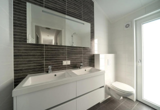 Baño moderno con doble lavabo, espejo grande, inodoro suspendido y abundante luz natural de la puerta.