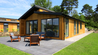 Lodge moderne en bois avec grandes baies vitrées, terrasse aménagée, entouré de pelouse et d’arbres verts.