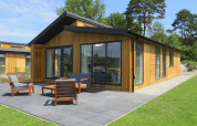 Moderne træhytte med store vinduer, terrasse og havemøbler, omgivet af grøn græsplæne og træer.