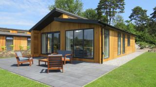 Moderne træhytte med store vinduer, terrasse og havemøbler, omgivet af grøn græsplæne og træer.