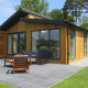 Moderne træhytte med store vinduer, terrasse og havemøbler, omgivet af grøn græsplæne og træer.
