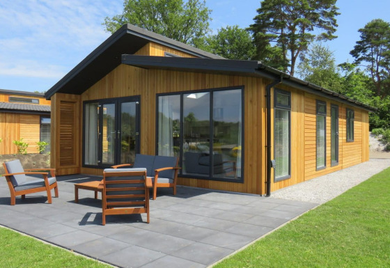Moderne houten lodge met ruime ramen en terras, tuinmeubilair en omgeven door groen gras en bomen.
