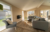 Salon lumineux et moderne dans un lodge avec grandes fenêtres, canapé gris et vue sur l’extérieur.