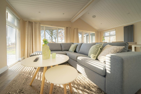 Helle Lodge-Wohnzimmer mit grauem Ecksofa, runden Tischen, großen Fenstern und gemütlicher Atmosphäre.
