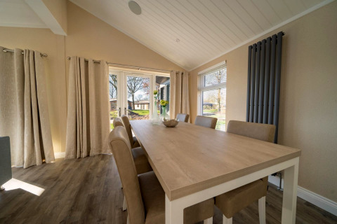 Salle à manger d’un lodge avec table en bois clair, six chaises, grandes fenêtres et rideaux beiges.