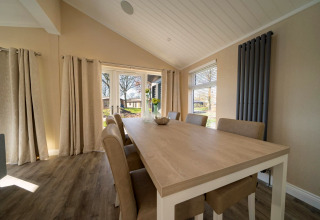 Salle à manger d’un lodge avec table en bois clair, six chaises, grandes fenêtres et rideaux beiges.