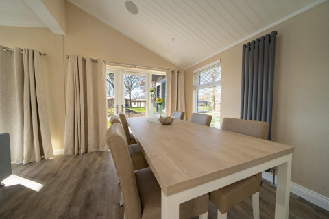 Comedor en una casa de campo con mesa de madera clara, seis sillas y grandes ventanales con cortinas beige.