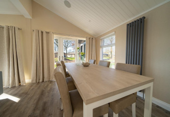 Comedor en una casa de campo con mesa de madera clara, seis sillas y grandes ventanales con cortinas beige.