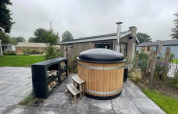 Wooden hot tub with black cover and stacked firewood at Hackfort Hottub 6, Zuiderzee, Netherlands, outdoors.