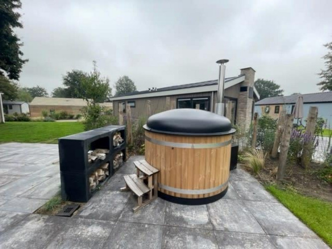 Wooden hot tub with black cover and stacked firewood at Hackfort Hottub 6, Zuiderzee, Netherlands, outdoors.