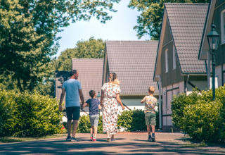 Una familia camina junto a modernas cabañas de glamping rodeadas de arbustos y árboles en un día soleado.