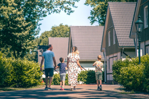En familie går blandt moderne glamping-hytter omgivet af grønne buske og træer på en solrig dag.