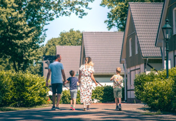 Een gezin wandelt langs moderne glampinghuisjes, omringd door groen en struiken op een zonnige dag.