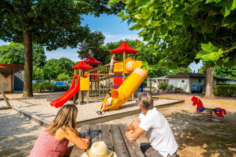 Des parents regardent leurs enfants jouer sur une aire de jeux colorée d'un site de glamping en été.