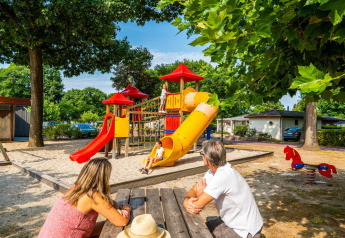 Familie nyder en solrig dag ved legepladsen på et glampingsted, mens børn leger på rutsjebaner.