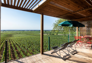 Terraza de madera con tumbonas y sombrilla, con vistas a viñedos verdes en Huttopia Meursault, Francia.