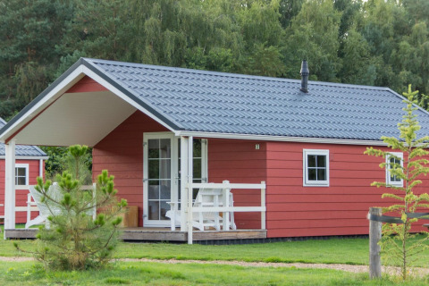 Boslodge 4, rood huisje met grijs dak en witte veranda, gelegen tussen groen en bossige bomen.