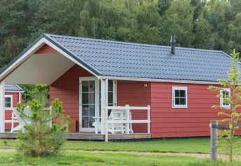 Boslodge 4, eine rote Hütte mit grauem Dach und Veranda, liegt inmitten von Bäumen und Grünflächen.