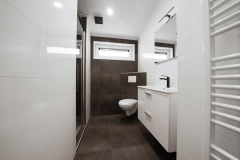 Baño moderno en un lodge con inodoro suspendido, ducha, ventanas y armario blanco debajo del lavabo.