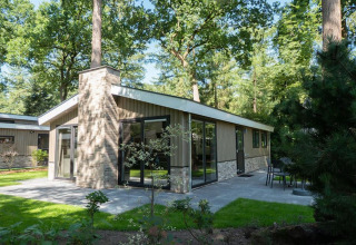 Moderne lodge met grote ramen en terras, midden in het groen en omgeven door bomen in een bosrijke omgeving.