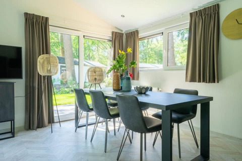 Eethoek in een lodge met een zwarte tafel, zes stoelen, grote ramen en een uitzicht op het groene buiten.