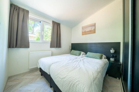 Dormitorio luminoso en una cabaña con dos camas individuales, cojines verdes y ventana con cortinas grises.