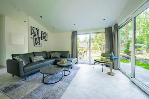 Sala moderna en una cabaña con ventanales, sofá gris, mesas de centro, sillón y vista al jardín exterior.