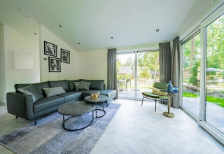 Sala moderna en una cabaña con ventanales, sofá gris, mesas de centro, sillón y vista al jardín exterior.