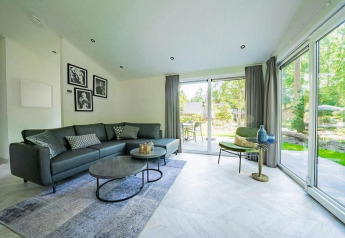 Sala moderna en una cabaña con ventanales, sofá gris, mesas de centro, sillón y vista al jardín exterior.