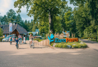 Una familia pasea en bicicleta junto al letrero de entrada de EuroParcs rodeado de naturaleza.
