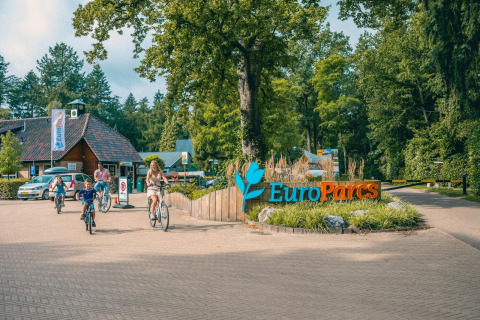 En familie cykler forbi indgangen til EuroParcs glampingsted omgivet af grønne træer og natur.