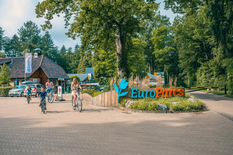 Een gezin fietst langs het EuroParcs-bord bij de ingang van een glamping, omgeven door bomen.