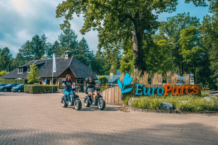 Two people on e-scooters at the entrance of EuroParcs glamping accommodation, surrounded by trees.