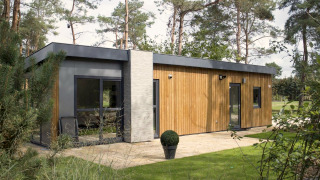 Lodge moderne en bois et béton niché dans une forêt, avec de grandes fenêtres et une pelouse bien entretenue.