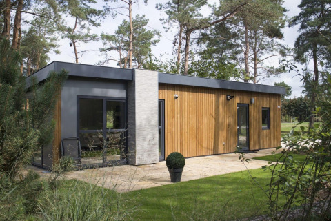 Modern wooden and concrete lodge surrounded by forest, featuring large windows and a neat green lawn.