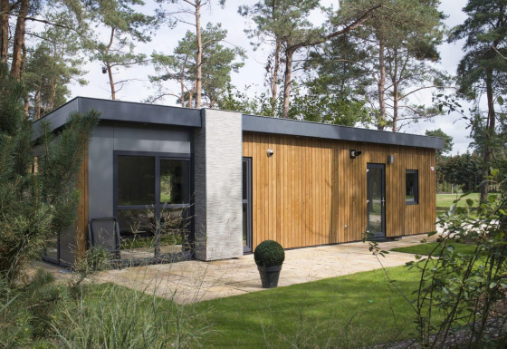 Moderne lodge i træ og beton beliggende i en frodig skov med store vinduer og velplejet græsplæne.