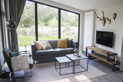 Modern glamping living room with large windows, a black sofa, nature view, and decorative antlers on the wall.