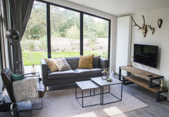 Moderne glamping woonkamer met grote ramen, zwarte zetel, natuurzicht en decoratieve gewei aan de muur.