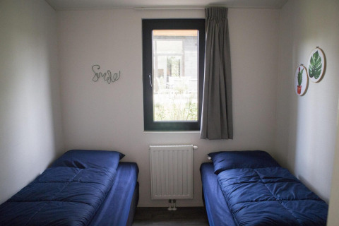 Habitación de glamping con dos camas individuales, ropa de cama azul y una ventana central con cortina gris.