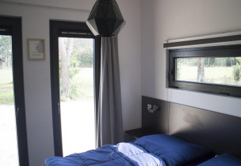 Moderne glamping accommodatie met grote ramen, blauw dekbed en uitzicht op de omringende natuur.