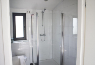 Baño moderno en alojamiento glamping con ducha de cristal, azulejos blancos y ventana luminosa.