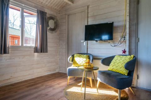 Sala luminosa con dos sillas modernas, cojines amarillos, TV en la pared y ventana en Stuga 4, De Utrechtse Heuvelrug.