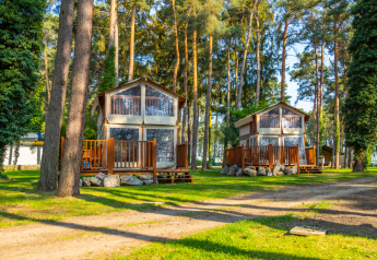 Twee Air Lodge chalets met houten terrassen in een zonnige bosomgeving, omringd door hoge pijnbomen en gras.