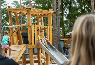Niños juegan en una estructura de parque de madera con tobogán junto a una cabaña en el bosque, adultos observan.