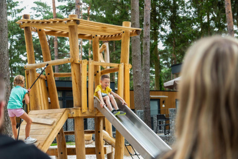 Des enfants jouent sur une aire de jeux en bois avec toboggan près d’un lodge en forêt, des adultes regardent.